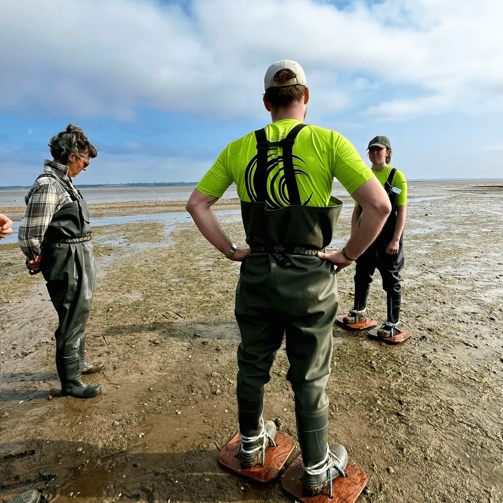 Three members of Project Seagrass staff are standing on a beach in Essex as part of May fieldwork. They are wearing mud shoes.