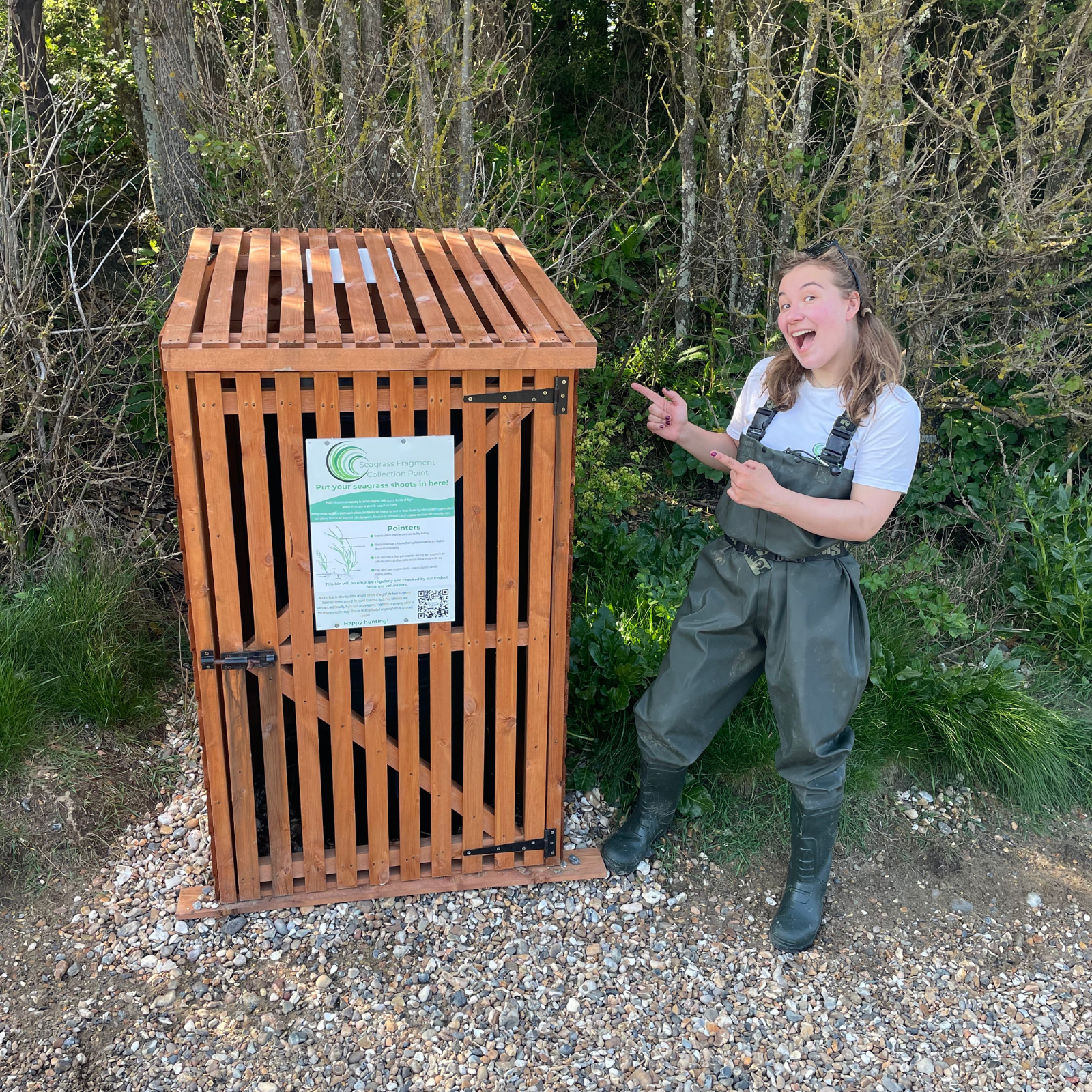 Becky is wearing waders and standing pointing at a seagrass fragment bin